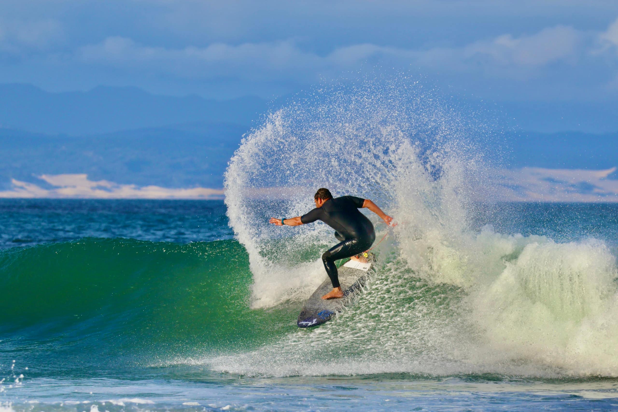 A surfer carving a powerful turn on a green wave, throwing a wide fan of spray, with a mountain coastline behind